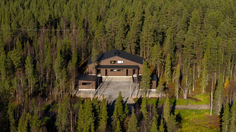 Villa Löhö seen from a distance, a small dark building in an expanse of autumn forest.