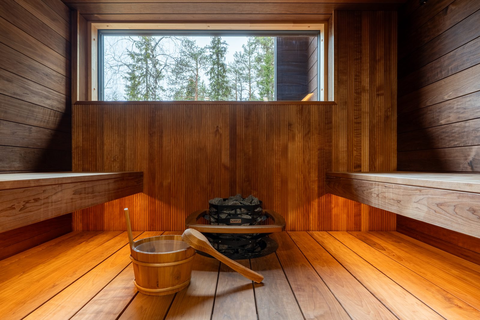 Inside the sauna: thermo-aspen benches on two levels, a bucket and ladle on the floor, and a window at bench height onto the forest.